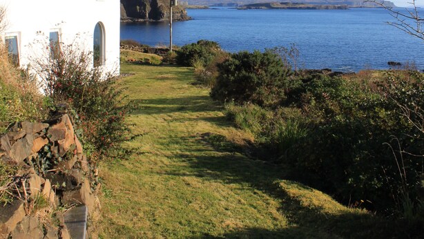 A white house with a black roof overlooking a calm body of water and distant hills.