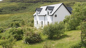 A white building with a sloped roof, surrounded by lush greenery and hills in the background.