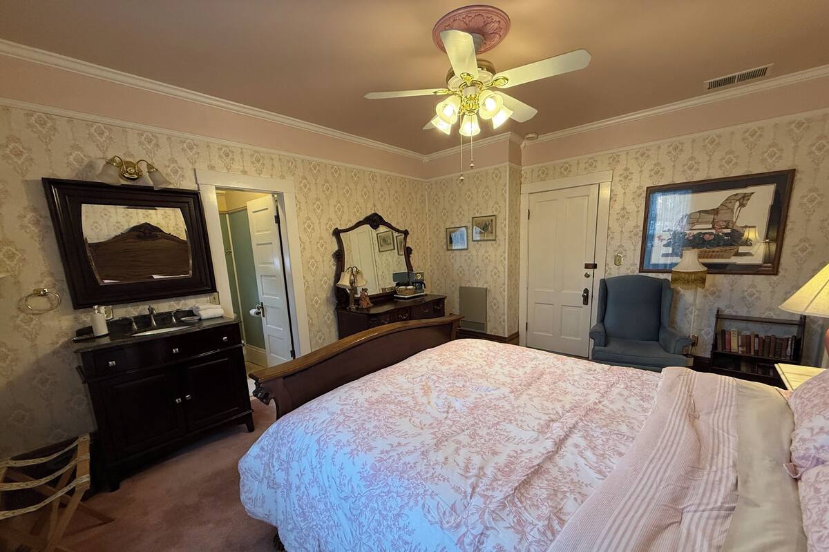 Ein Schlafzimmer mit einem großen Bett, einem Schminktisch, einem Kleiderschrank, einem Deckenventilator und einem Bücherregal.