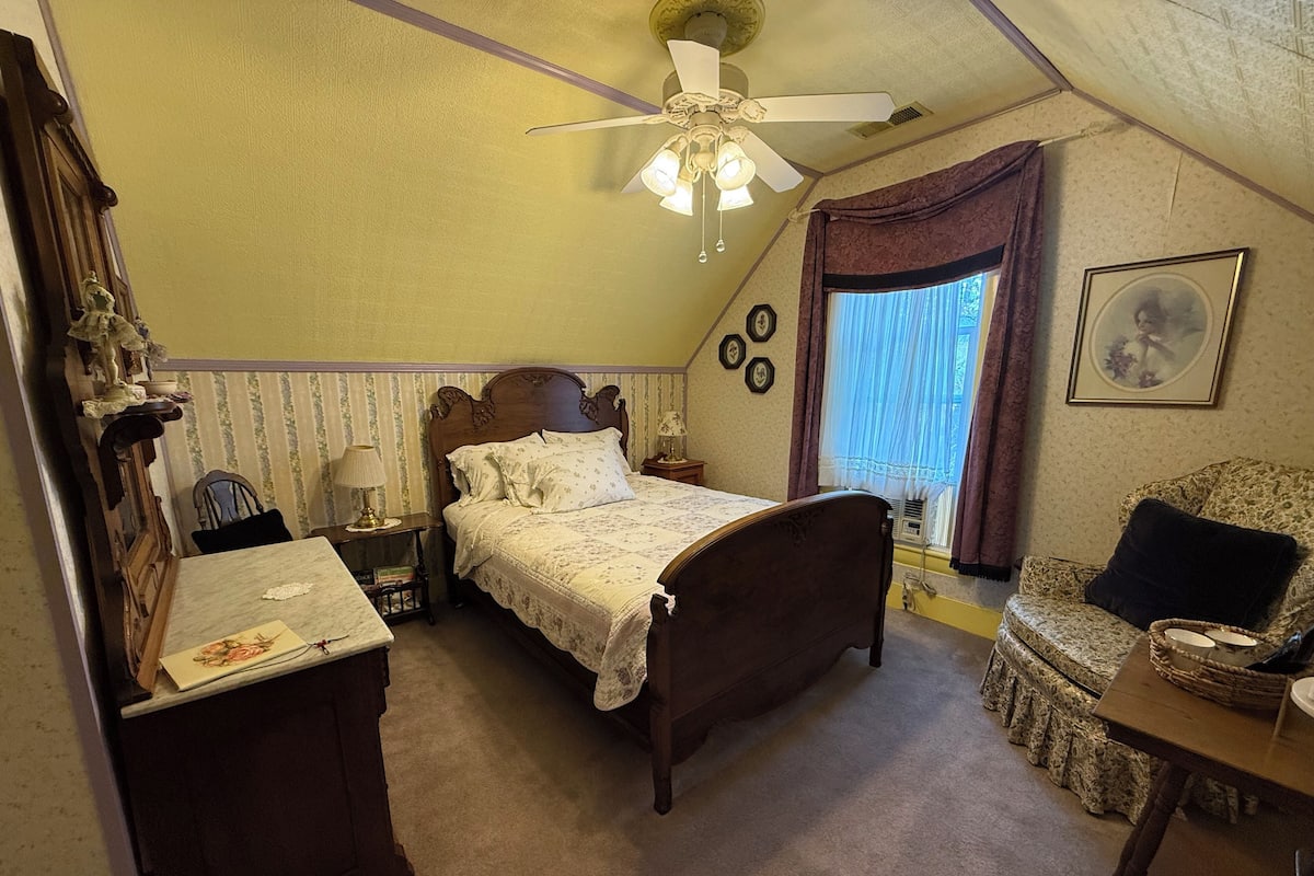 Ein Schlafzimmer mit Bett, Schreibtisch, Stuhl, Deckenventilator und einem Fenster mit Vorhängen.