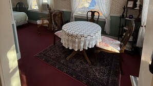 Een kamer met een ronde tafel bedekt met een witte kanttafelkleed, twee houten stoelen, een kamerkleed met een patroon, een plafondventilator en een raam met kanten gordijnen.