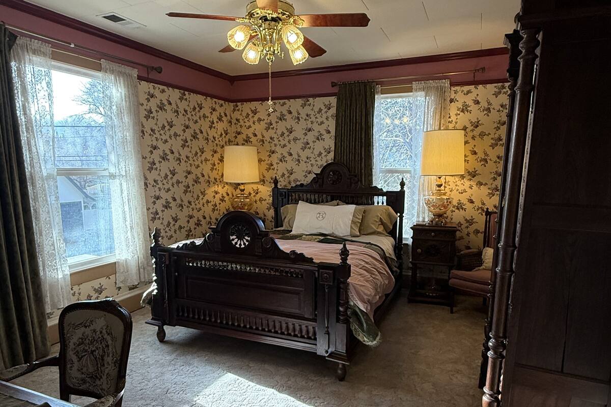 Ein Schlafzimmer mit einem großen Bett, einem Schreibtisch und einem Stuhl. Es gibt einen Deckenventilator und zwei Fenster mit Vorhängen.