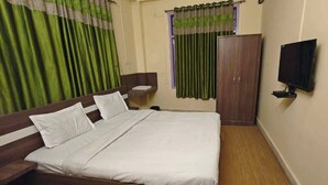 Uma cama bem arrumada com lençóis brancos, cabeceira de madeira, uma pequena mesa de cabeceira com abajur, um guarda-roupa e uma televisão na parede.