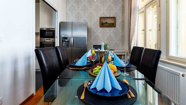 Un coin repas avec une table en verre, des chaises noires et un lustre. On aperçoit au fond une cuisine équipée d’un réfrigérateur et d’un four.
