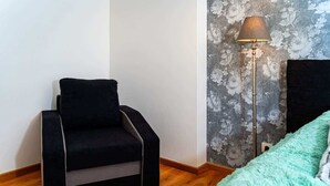Un petit fauteuil noir avec un coussin gris, un lampadaire avec un abat-jour gris et un plaid turquoise sur un lit.