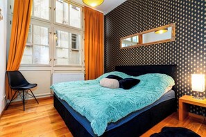 A bedroom with a bed, a chair, a nightstand, a mirror, and a window with curtains. - Golden Dog Kingdom (Hlavní mesto Praha)