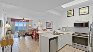 A modern kitchen with white cabinetry, a built-in dishwasher, and a stainless steel oven. There is a dining area with a table and chairs, and a view of the ocean through large windows. - Windemere 1206 (Pensacola)
