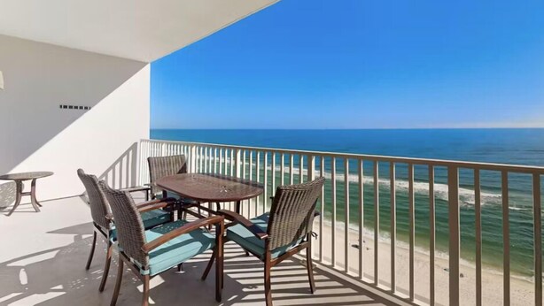 A balcony with a table and chairs, overlooking a beach and ocean. - Windemere 1206 (Pensacola)