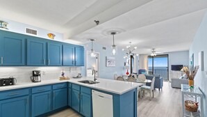 A modern kitchen with blue cabinets, white countertops, and a dishwasher. There is a dining area with a table and chairs, and a view of the ocean through large windows. - Shoalwater 1005 (Orange Beach)
