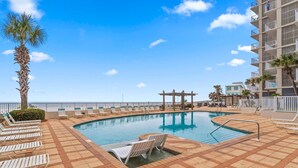 A pool area with lounge chairs, a palm tree, and a building with balconies. - Shoalwater 1005 (Orange Beach)