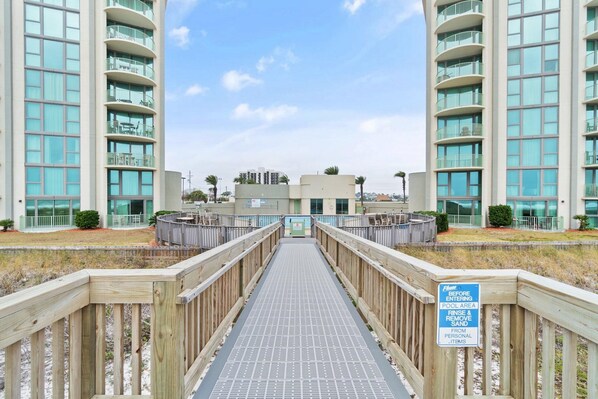 A wooden boardwalk leading to a modern building with balconies and a sign indicating a pool area. - Perdido Towers East 503 (Pensacola)
