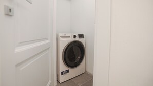 Ein kompakter, weißer Waschraum mit einer Frontlader-Waschmaschine, einer kleinen Ablage darüber und einer Tür links.