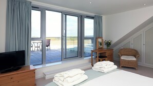 Ein Schlafzimmer mit einer großen Schiebeglasdoor zum Balkon, einem Fernseher auf einer Holzkommode, einem Bett mit weißen Leinen, einem Korbstuhl und einem kleinen Holztisch mit Spiegel und Blumen.