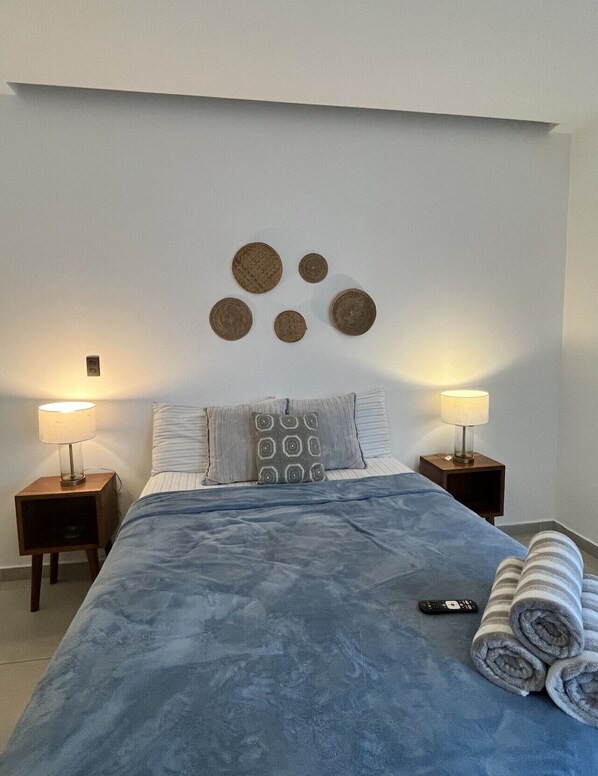 A neatly made bed with a blue blanket, two bedside lamps, and a remote control on the bed.