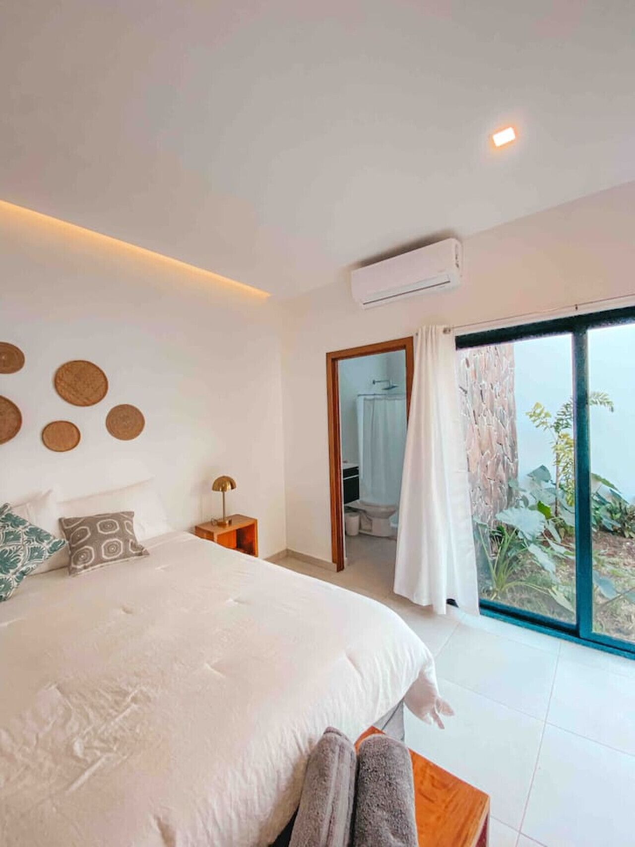 A bedroom with a large bed, a bedside table, a nightstand, an air conditioning unit, and a view of the outdoors through a glass door. - Villa Uma • 50m to Zonte wave • 2Bdr (El Zonte)