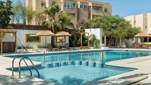 Una piscina con solárium, sillas de descanso y un edificio con balcones al fondo.