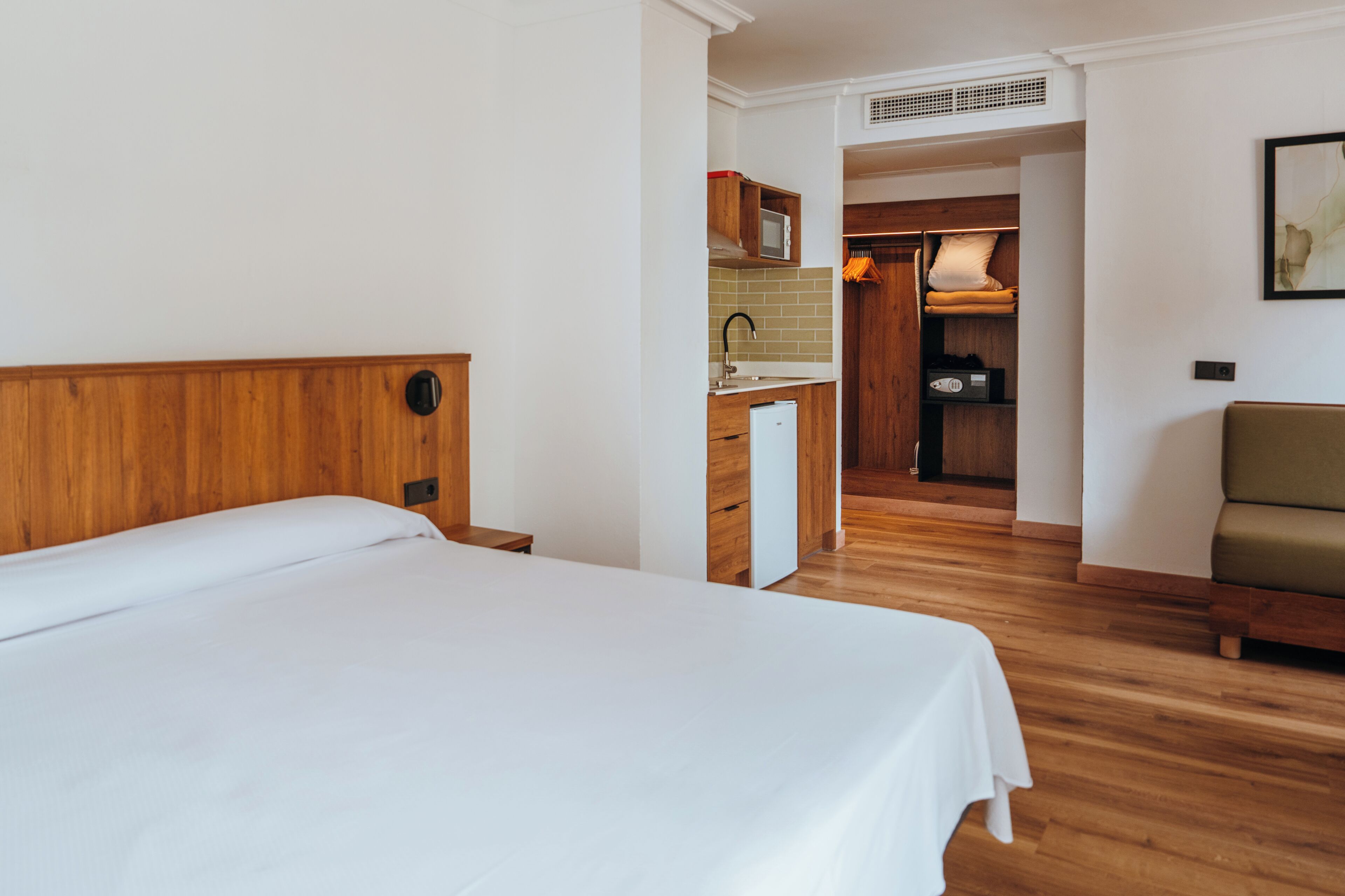 Habitación de hotel con cabecera de madera, cama, kitchenette y zona de estar.