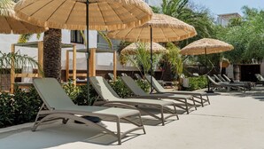 Zona junto a la piscina con sillones de descanso y sombrillas de paja, rodeada de palmeras y un edificio con fachada blanca.