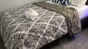 A neatly made bed with patterned bedding and a folded towel, a dark upholstered headboard, and a window with blinds. - Luxurious 2 Bdrm Suite ~ North Edmonton (3min to Anthony Henday/Manning Dr) (Edmonton)
