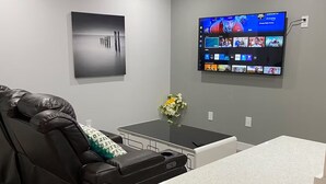 A modern living room with a leather sofa, a small table with a vase of flowers, and a mounted TV displaying various channels. - Luxurious 2 Bdrm Suite ~ North Edmonton (3min to Anthony Henday/Manning Dr) (Edmonton)