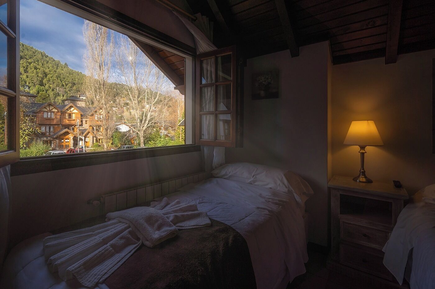 Un dormitorio con una cama, una mesita de noche con una lámpara, una ventana con vista a una casa y árboles, y un radiador debajo de la cama.