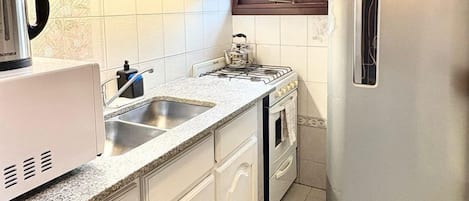Une cuisine avec des meubles blancs, un réfrigérateur en acier inoxydable, une cuisinière à gaz et une fenêtre qui laisse entrer la lumière naturelle.