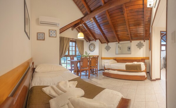 A hotel room with two beds, a wooden ceiling, a dining area with a table and chairs, and a clock on the wall.