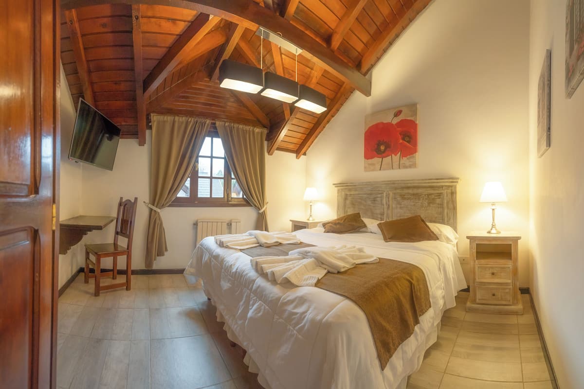 A bedroom with a large bed, wooden ceiling beams, a window with curtains, a mounted TV, and a bedside lamp.