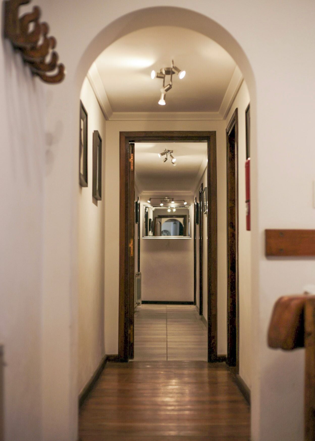 A narrow hallway with a wooden floor, a ceiling light fixture, and framed pictures on the walls.