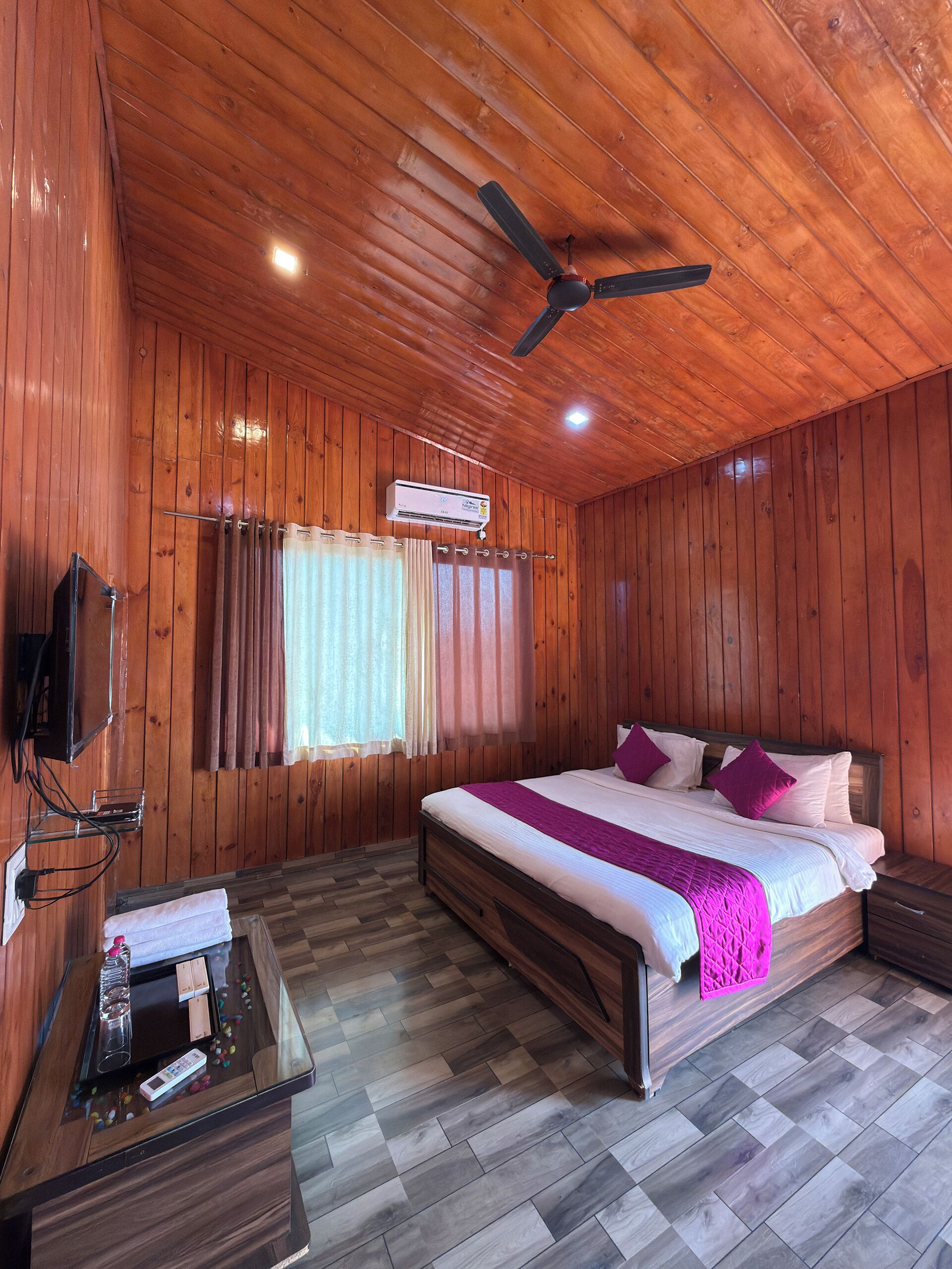 A wooden-paneled room with a bed, a ceiling fan, an air conditioning unit, a TV, and a bedside table with a remote control.