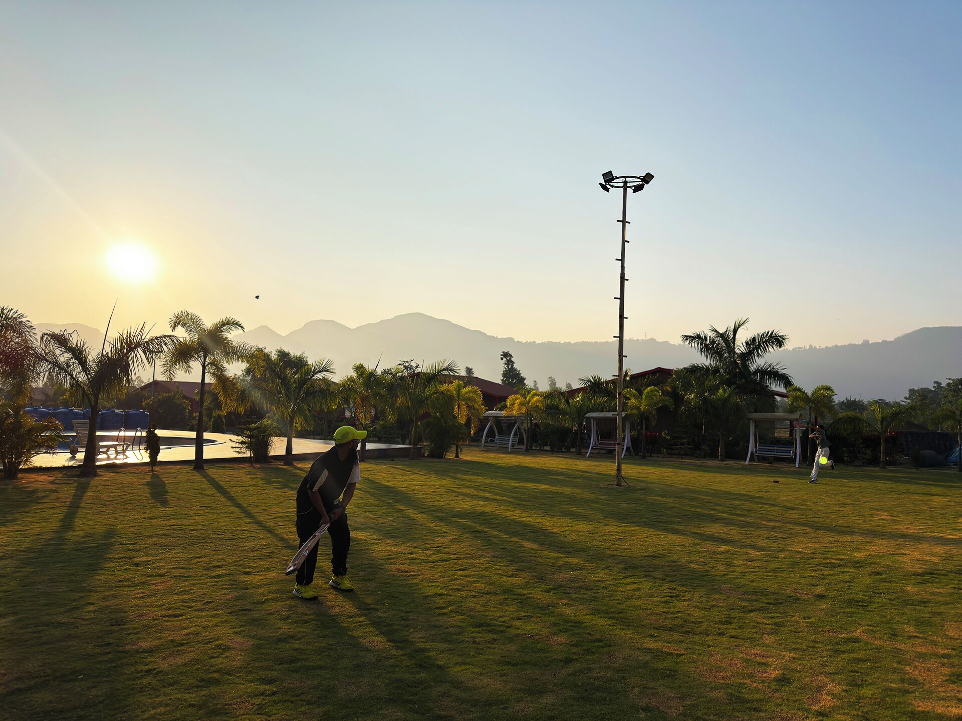 A grassy outdoor area with a person holding a frisbee, a light pole, and a clear sky with the sun low on the horizon.