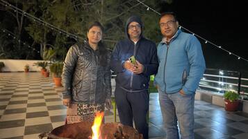 Tres personas cerca de una fogata en un área exterior a cuadros con plantas en macetas y luces colgantes.
