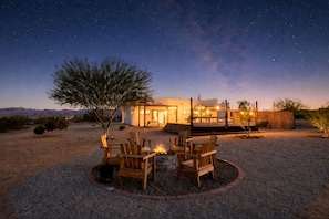 A desert-style building with a fire pit and chairs in the foreground, a deck with lights, and a starry night sky.