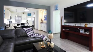 A modern living room with a leather sofa, a coffee table, and a TV on a wooden stand. There is a kitchen in the background with white cabinets and a dining area. - C-204 Enjoy Your Vacation In This Exquisite Executive Condominium! (Cabo San Lucas)