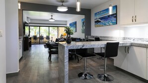 A modern kitchen with a marble countertop, black bar stools, and a large island. There are two pendant lights above the island and a painting on the wall. - C-204 Enjoy Your Vacation In This Exquisite Executive Condominium! (Cabo San Lucas)