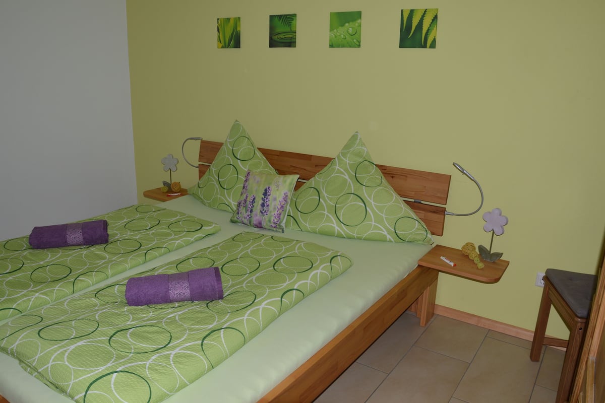 Cama doble con ropa de cama verde estampada y dos toallas moradas. Hay una mesita de noche de madera con una lámpara y una pequeña planta.