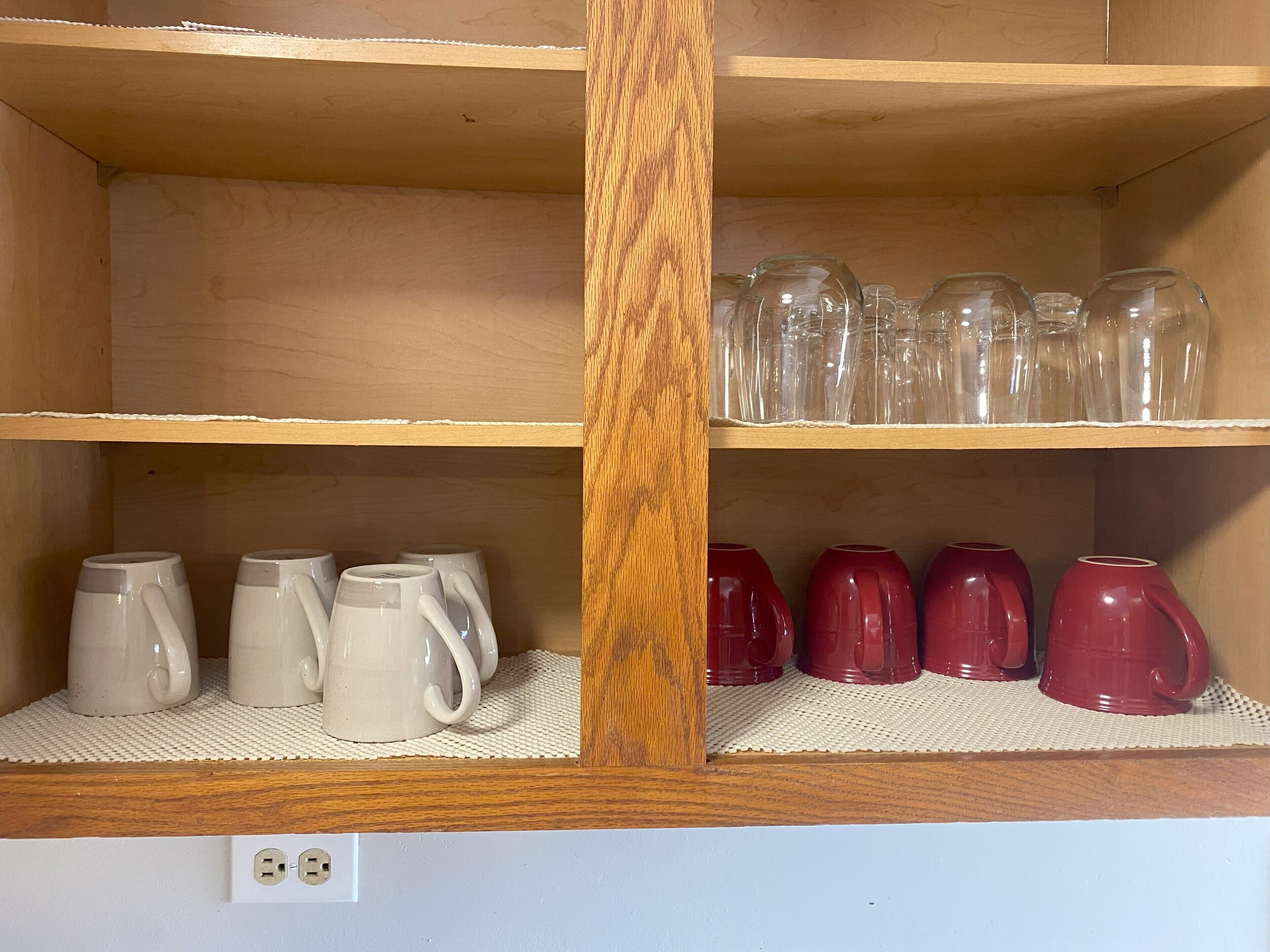 A wooden shelf unit with two levels, the top holding clear glass tumblers and the bottom holding white and red mugs.