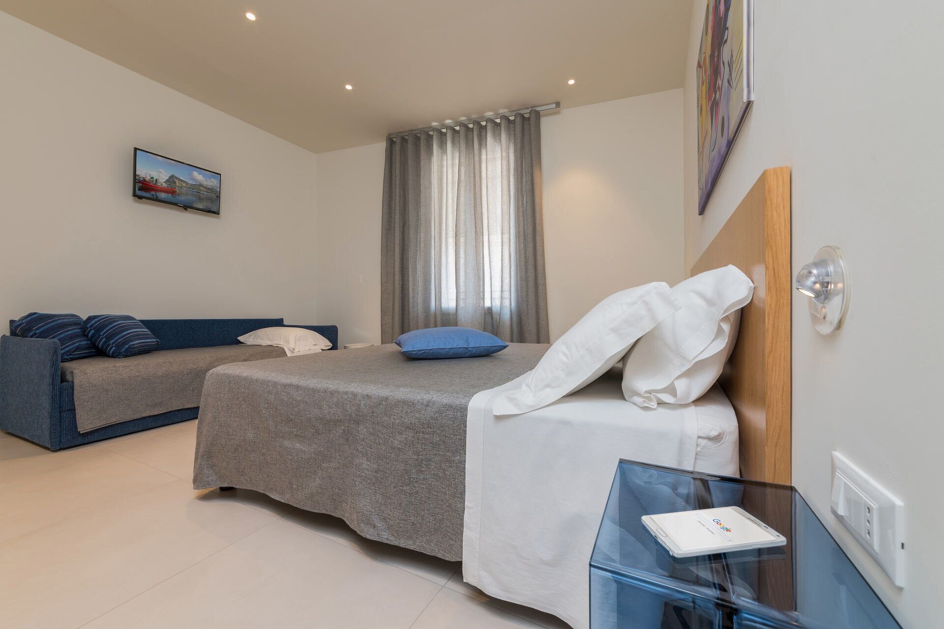 Habitación de hotel con cama, sofá, mesa de vidrio con una computadora portátil, un cuadro en la pared y ventana con cortinas.