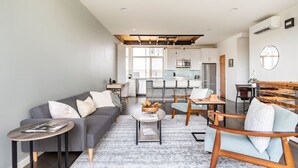 A modern living room with a grey sofa, a coffee table, and a wooden bench. There is a kitchen in the background with white cabinets and a dining area with a table and chairs.