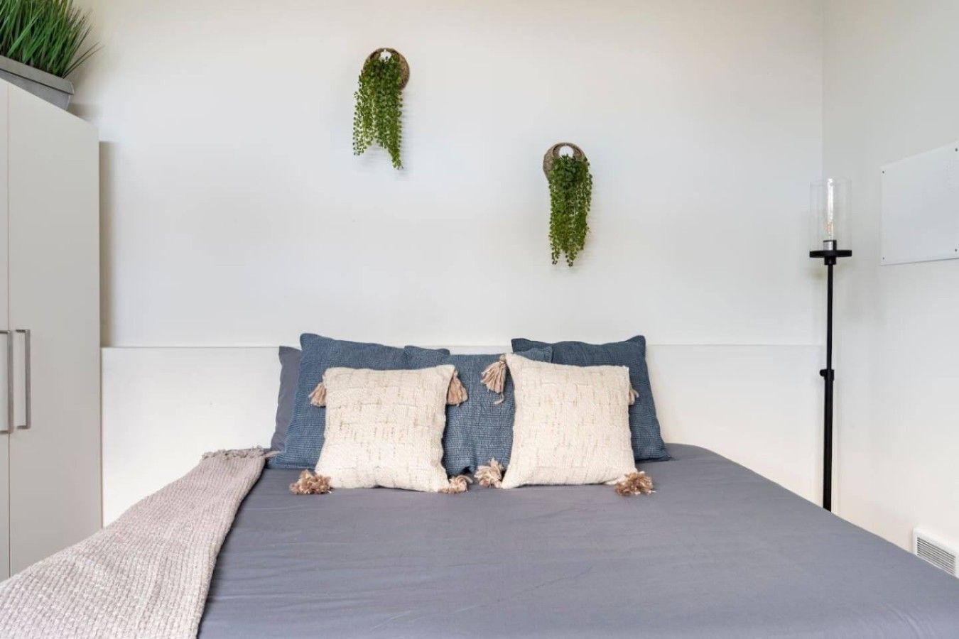 A bedroom with a bed, pillows, a bedside table, and two hanging plants.