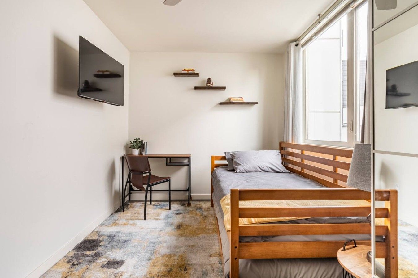 Une petite pièce, de taille modeste, comprenant un lit, un bureau et une chaise. Il y a trois étagères au mur et une fenêtre sur un des côtés.