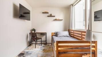 Une petite pièce, de taille modeste, comprenant un lit, un bureau et une chaise. Il y a trois étagères au mur et une fenêtre sur un des côtés.
