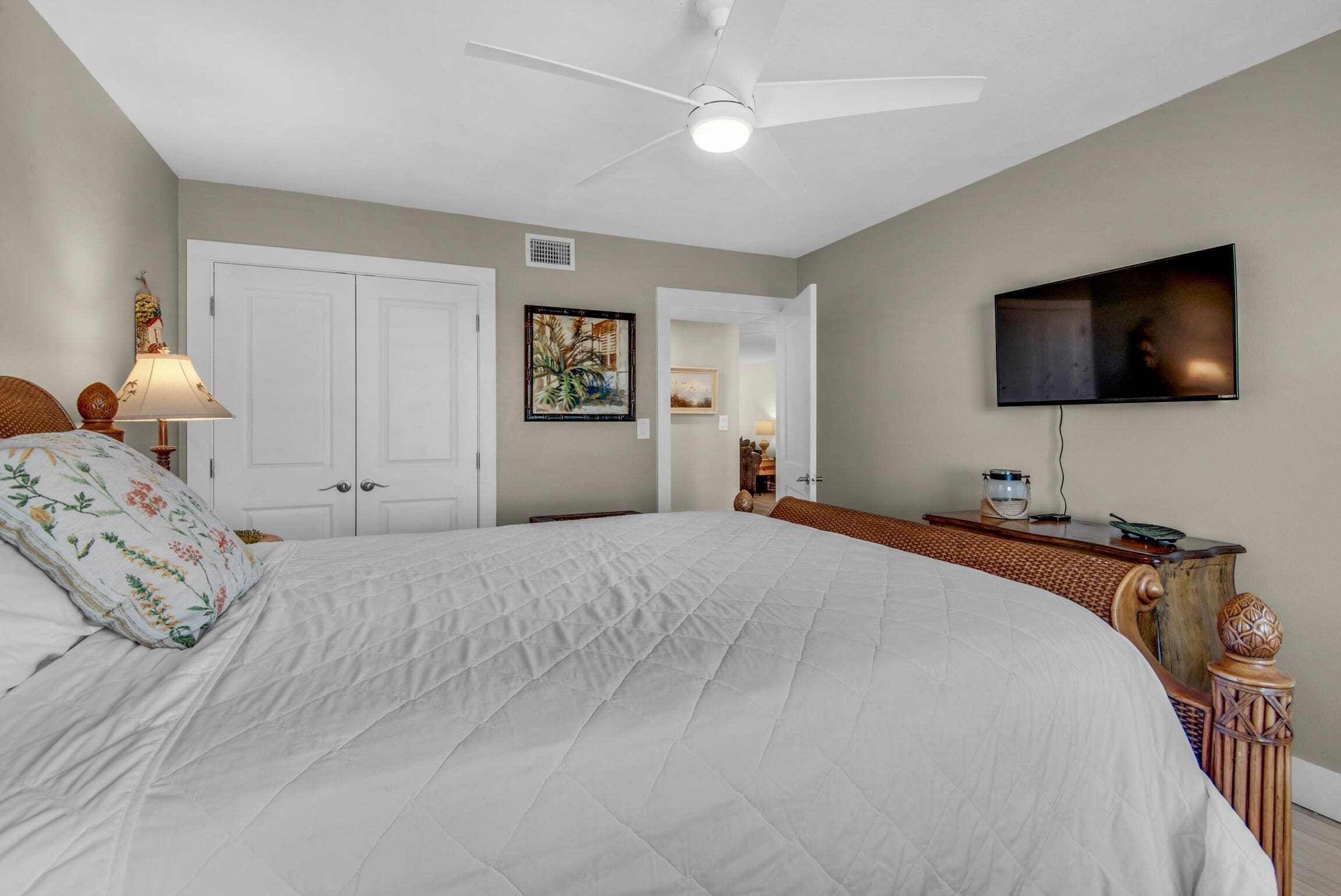 A bedroom with a large bed, a ceiling fan, a TV mounted on the wall, and a door leading to another room.