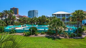 A pool area with a green fence, palm trees, and a building with a white roof in the background. - Deep Water Cove 103 (Pensacola Beach)