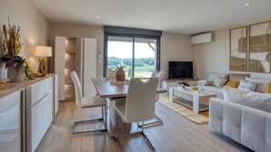 A modern living room with a sofa, coffee table, and TV. There is a dining area with a table and chairs. A large window provides a view of the outdoors. - L'Annexe Coté Castagne du Domaine des Hauts de Rouquette (Eymet)