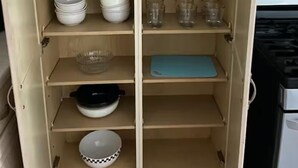 A kitchen with wooden cabinets, shelves with plates, bowls, cups, and a blue cutting board.
