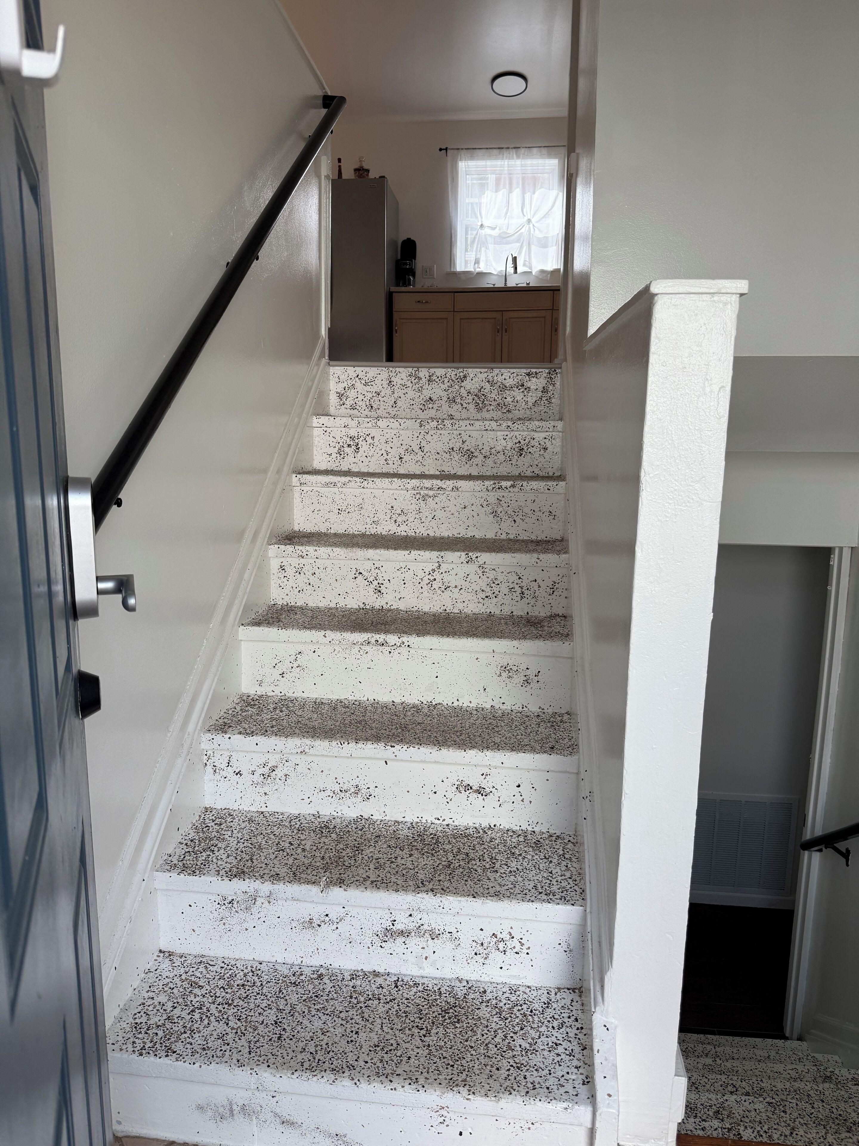 A staircase with white steps and black speckles, leading to a kitchen area with wooden cabinets and a window with curtains.