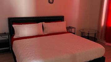 Una cama grande con cabecera negra y ropa de cama blanca, dos apliques de pared y una mesita de noche con una silla.