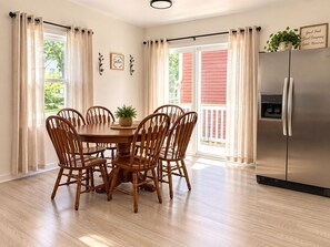 A dining area with a wooden table and chairs, a refrigerator, and a view of a red building through the windows.