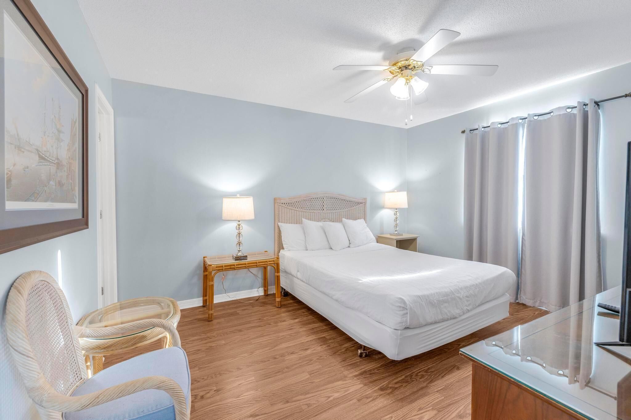 A bedroom with a bed, two bedside tables with lamps, a ceiling fan, a chair, and a glass table.
